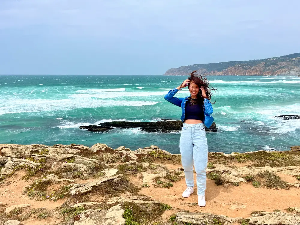 The blog's author standing on rocky sand by a teal ocean while the wind blows her hair.