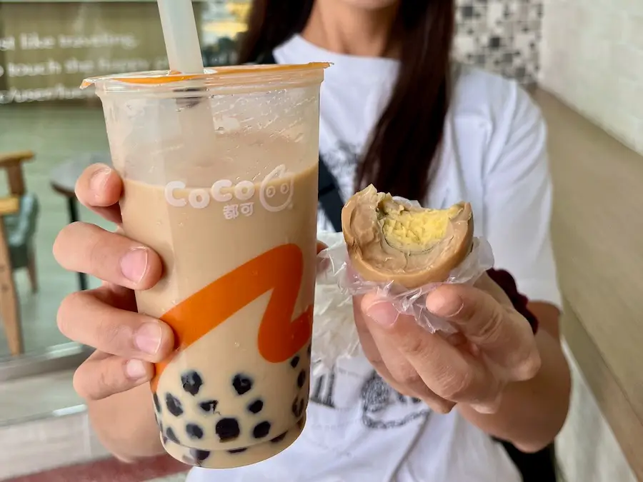 Woman holding up a milk boba tea and half-bitten preserved egg.