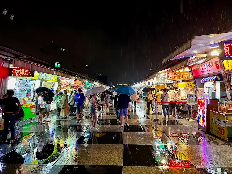 A night market with shops showing bright lights while people walk around.