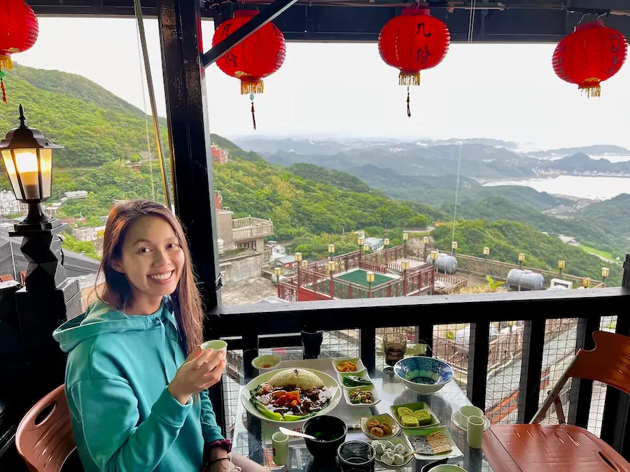 The blog author holding a small cup of tea sitting next to a view of mountains and red lanterns.