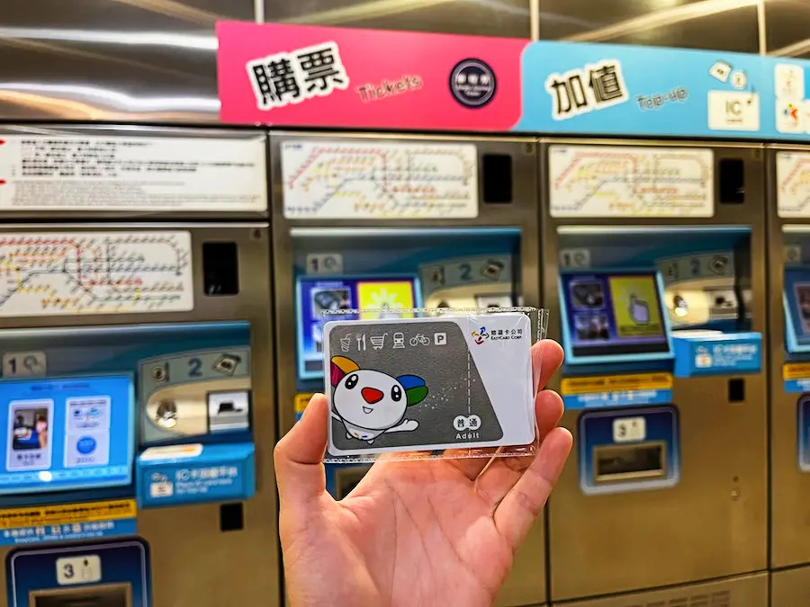 A hand holding a card with a cute cartoon on it, with metro kiosks in the background with Chinese writing on them.