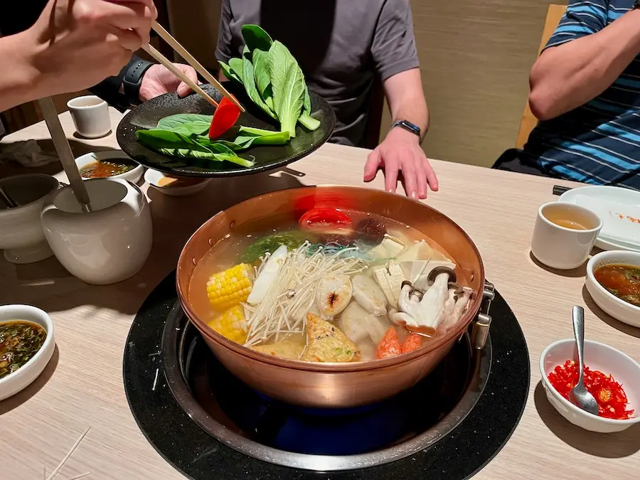 A pot with broth containing vegetables, mushrooms, corn, etc, over a stove on a dining table.