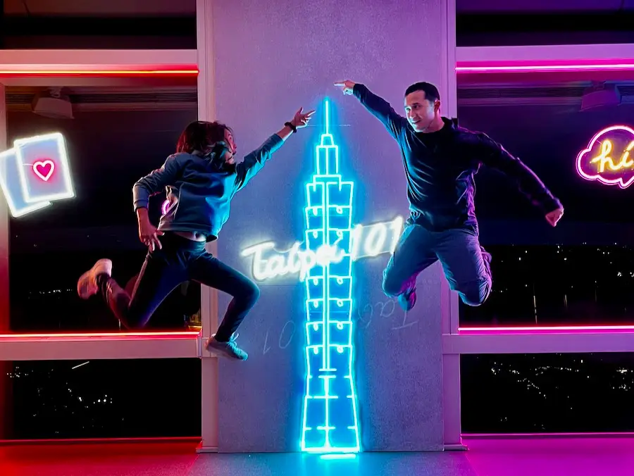 Two people jumping and posing in front of a light-up sign that says "Taipei 101".