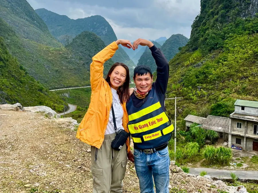 The blog author wearing a black, crossbody pouch while making a heart with a man that is wearing a vest that says "Ha Giang Epic Tour."
