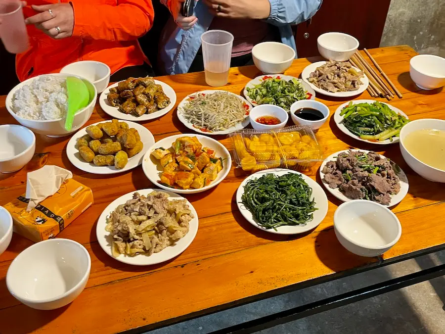 A wooden table with plates of cooked vegetables, noodles, meat, soup and rice.