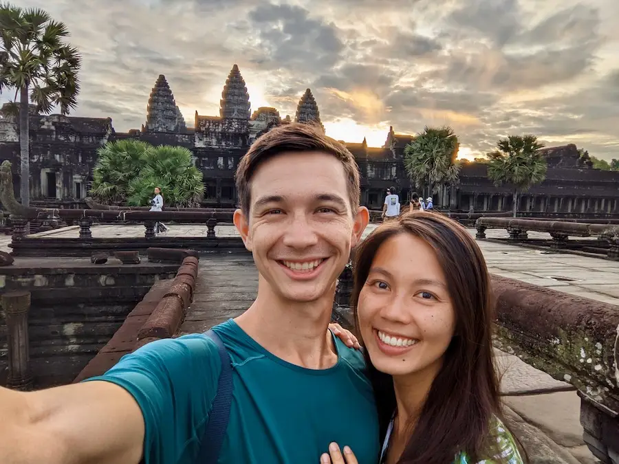 Two people take a selfie in front of Angkor Wat while the sun rises in the background.