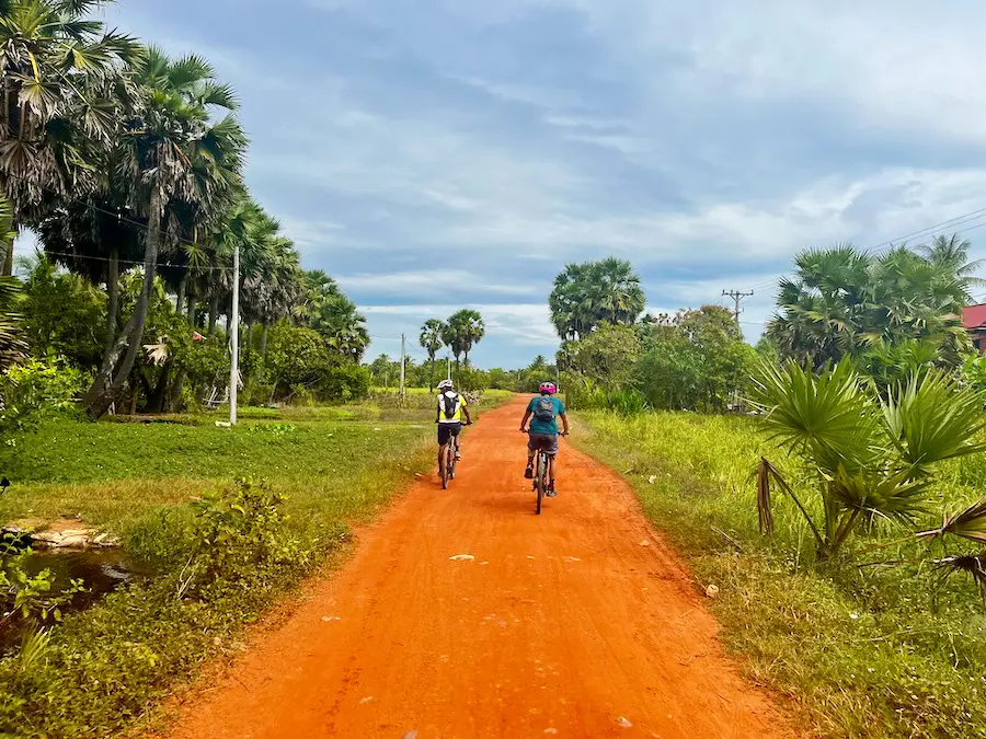 Two people riding bikes down a red-dirt road flanked by green trees.