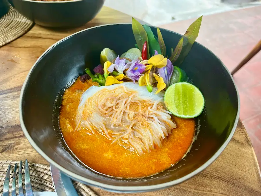Noodles in an orange soup with flowers and a lime adorning the side.