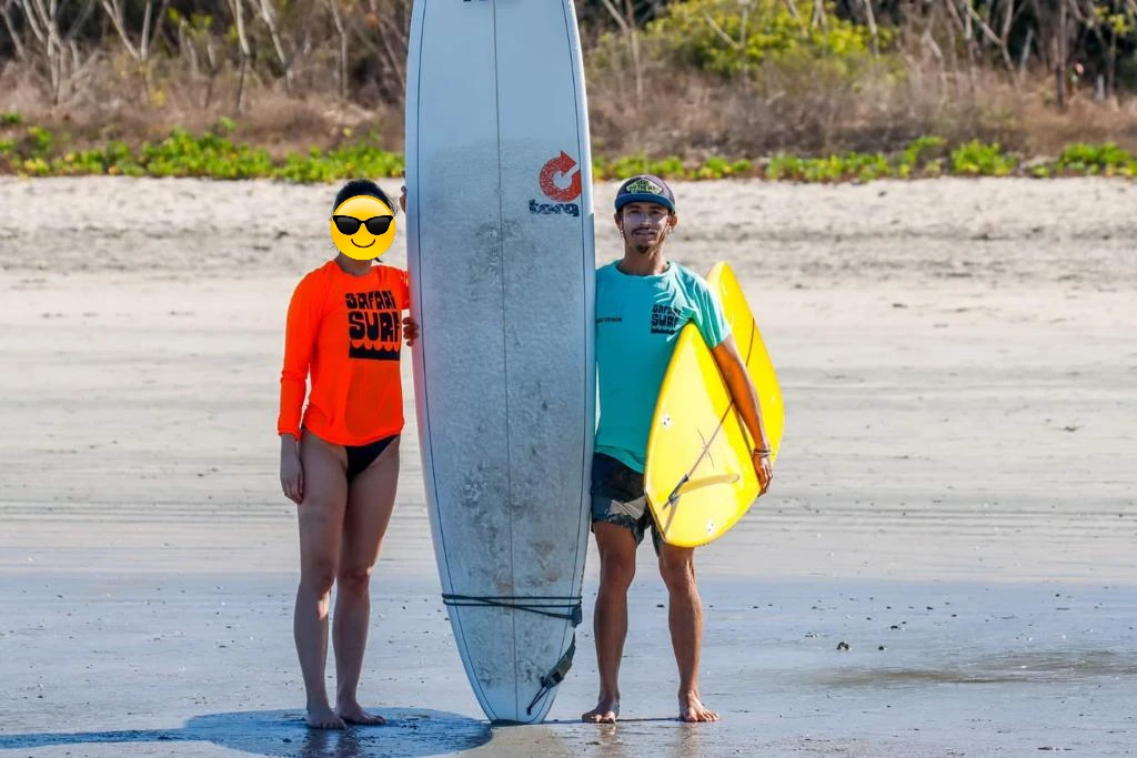 Two people standing next to an upright surfboard on sand.
