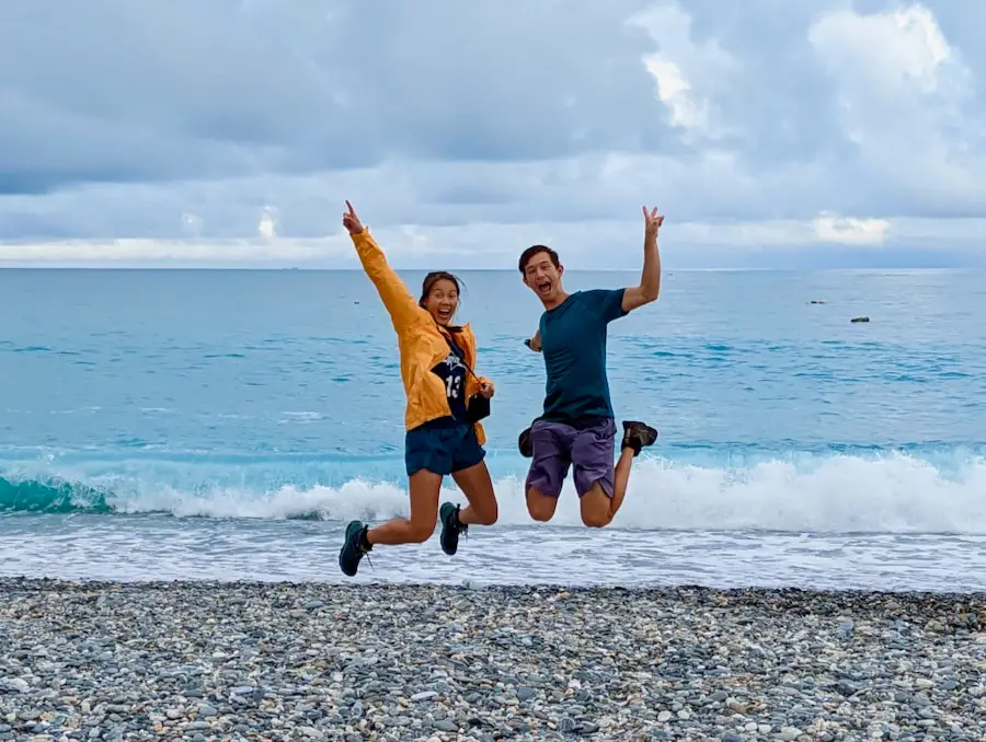 Two people jumping and making a pose in front of a blue ocean and pebbled beach.