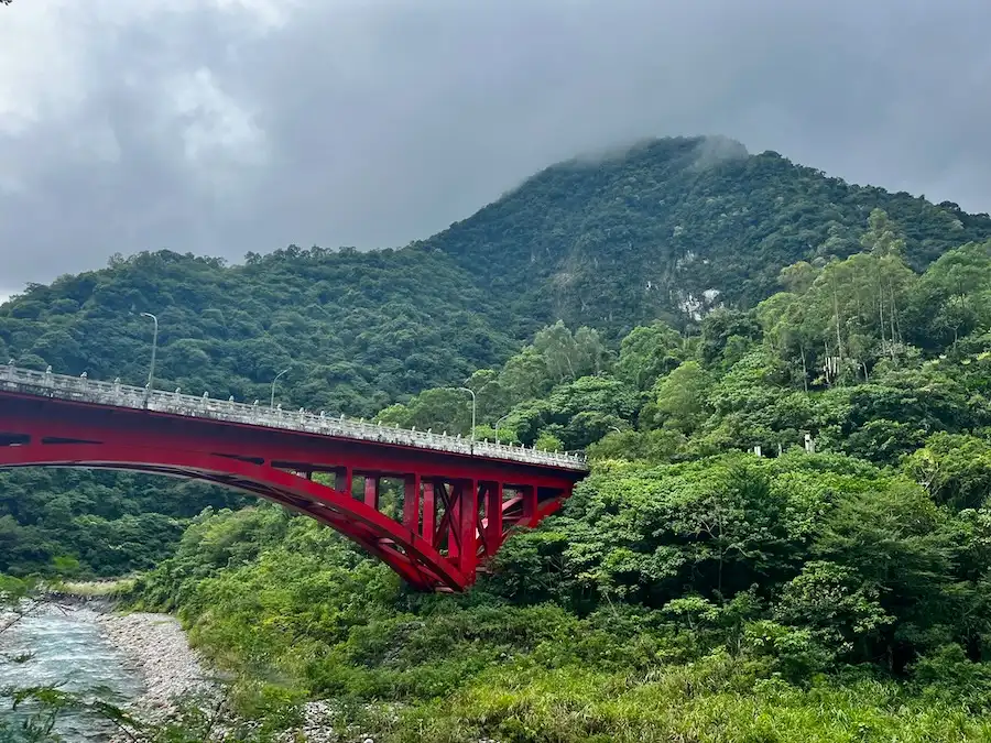 A red bridge that connects to a green, lush mountain with trees.