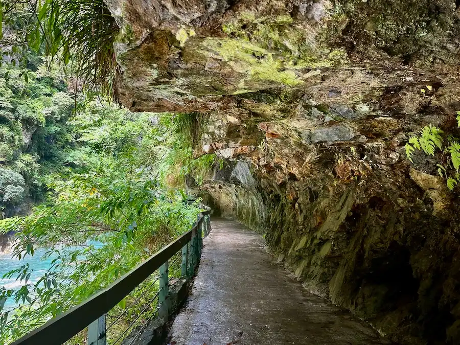 A carved marble mountain creating a partial tunnel for a walkway. A blue river is on the left with green plants.