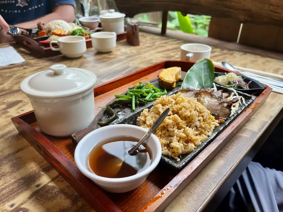 Rice, pork belly, vegetable and a soup on a wooden tray. All food items are separated and organized.