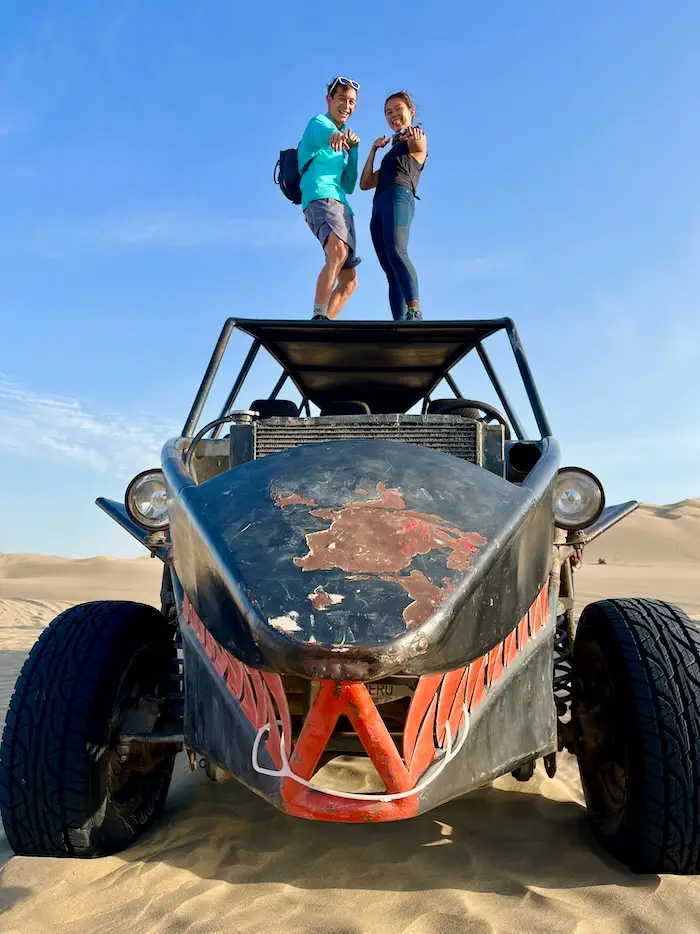 Two people standing on a roof of an open vehicle on sand.