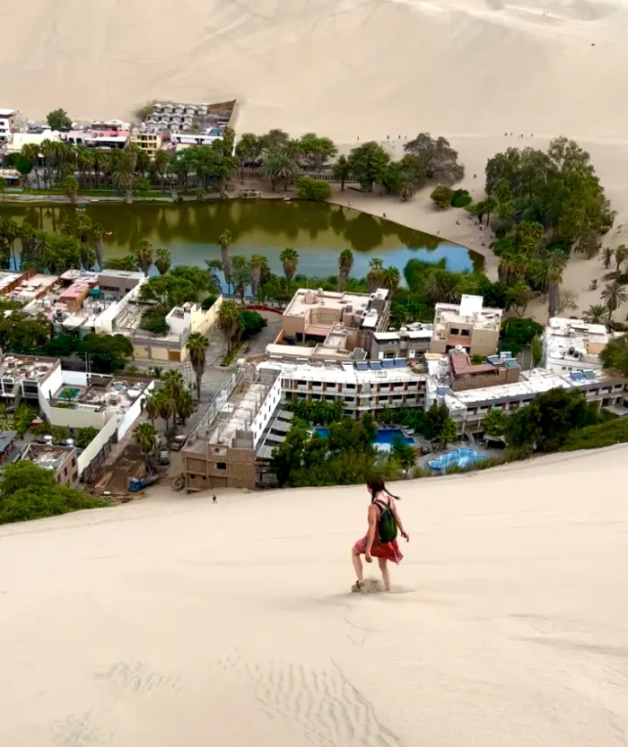A woman walking down a steep sand dune overlooking a village with trees.