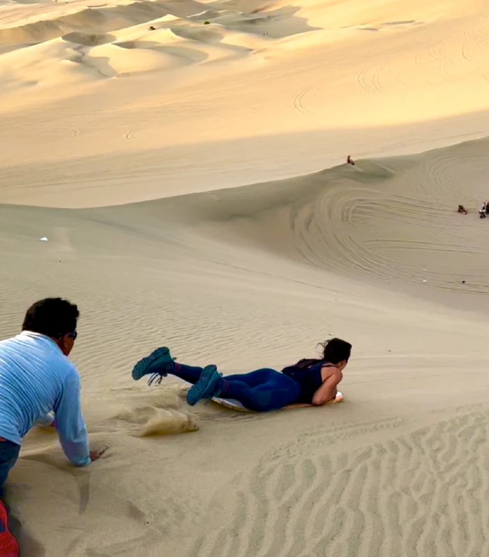 A woman on a board, laying on her stomach, about to slide down a sand dune.