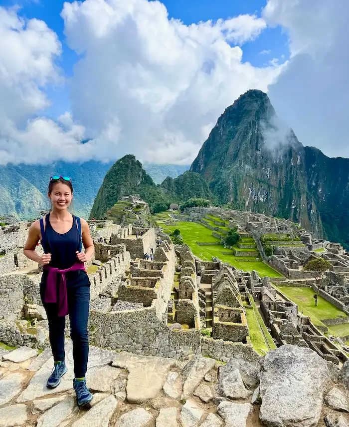 A woman standing on a ledge with Inca ruins and mountains in the background.