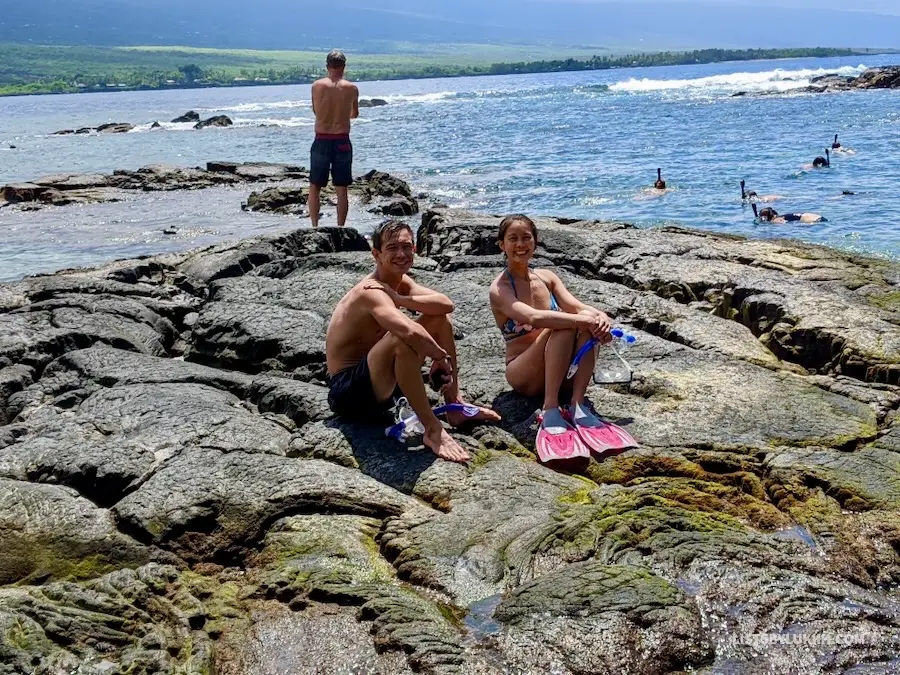 Two people with snorkel gears on sitting on a black rock near the ocean.