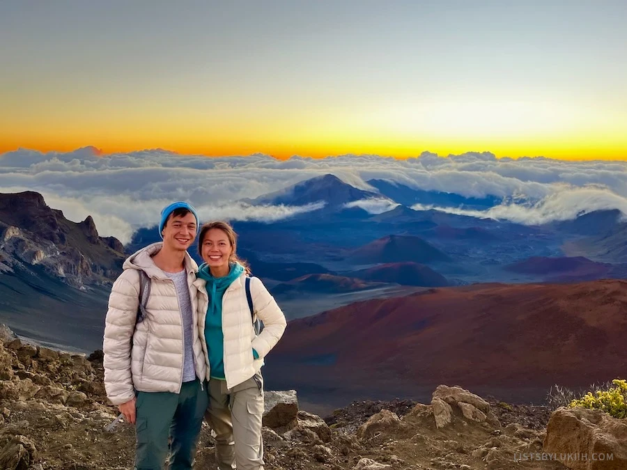Two people standing on a mountain with clouds and a sunrise in the background.