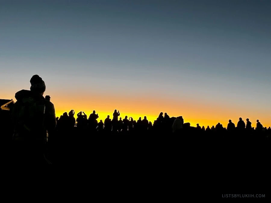 The silhouette of many people lined up and waiting for the sun to rise on a mountain.