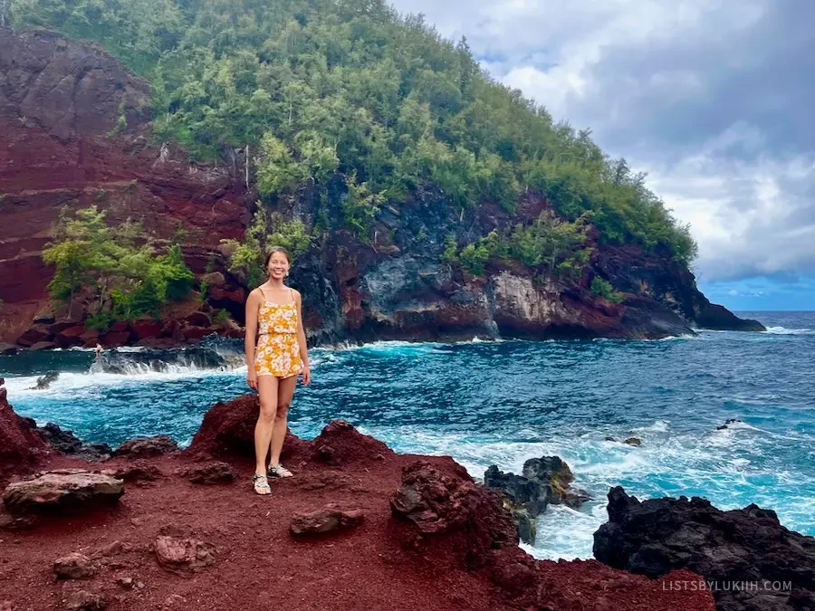 A woman standing on red sand with a very blue ocean in the background.