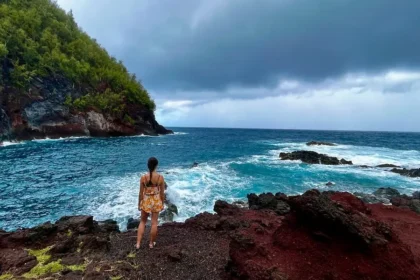 A woman standing on the cliff of red sand looking out at a turquoise ocean.