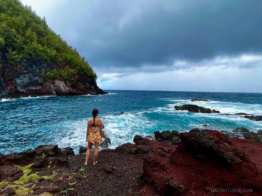 A woman standing on the cliff of red sand looking out at a turquoise ocean.