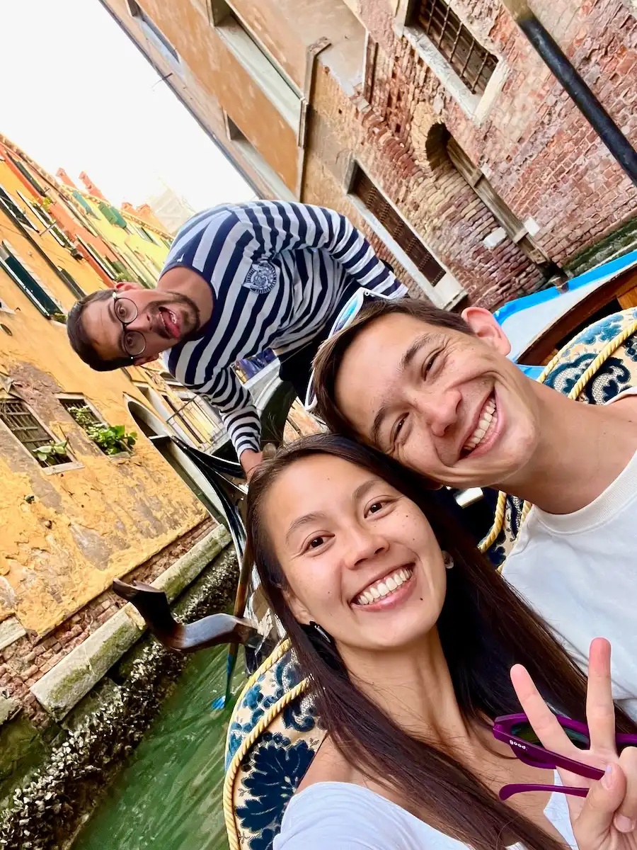 Three people taking a selfie on a boat in a small canal. One man is standing and wearing a striped shirt.