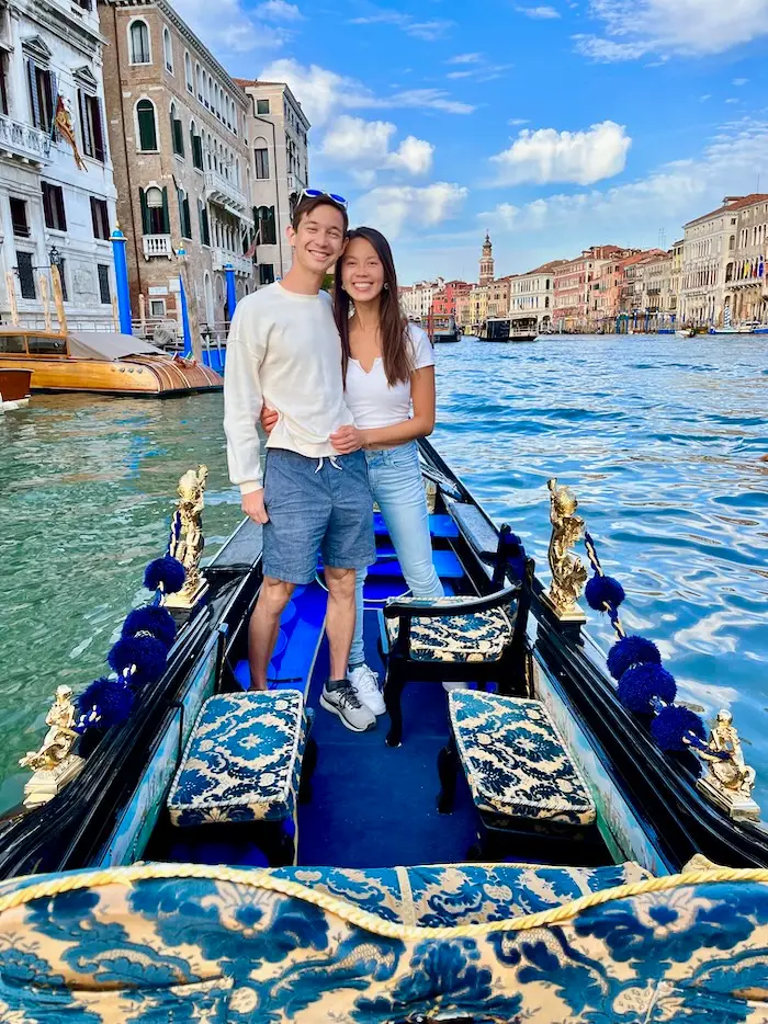 Two people standing on a blue boat in a big canal surrounded by colorful buildings.