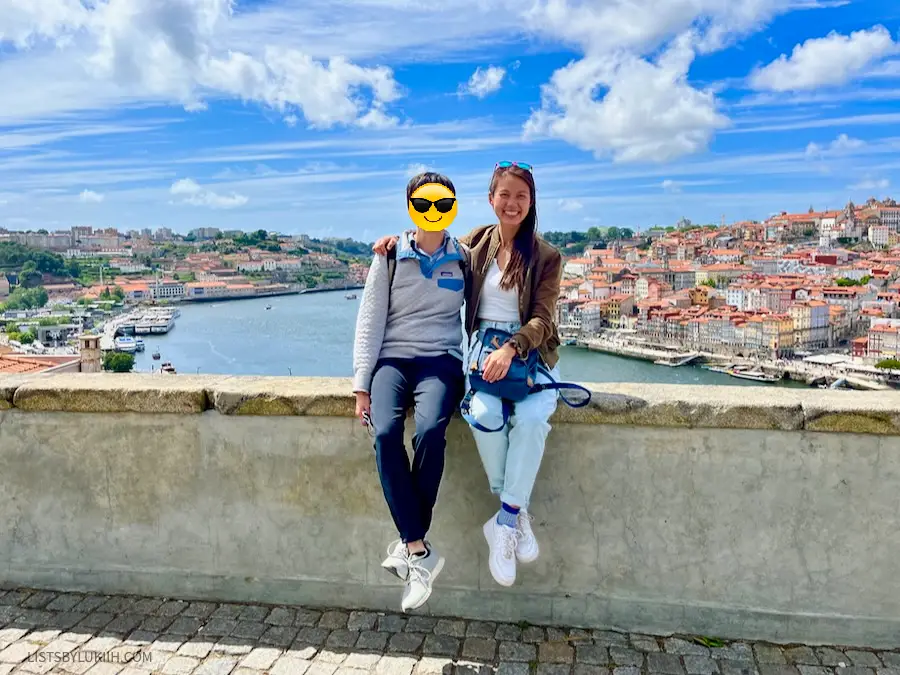 Two women sitting on a lookout ledge overlooking a body of water flanked by orange roof buildings.