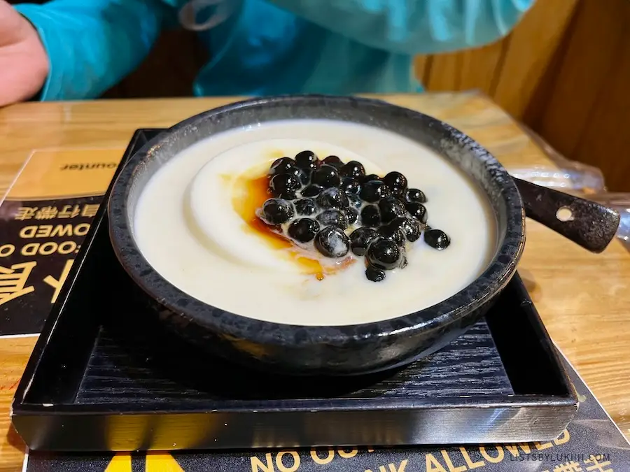 A black bowl contain milky white pudding and sugar-glazed tapioca pearls.