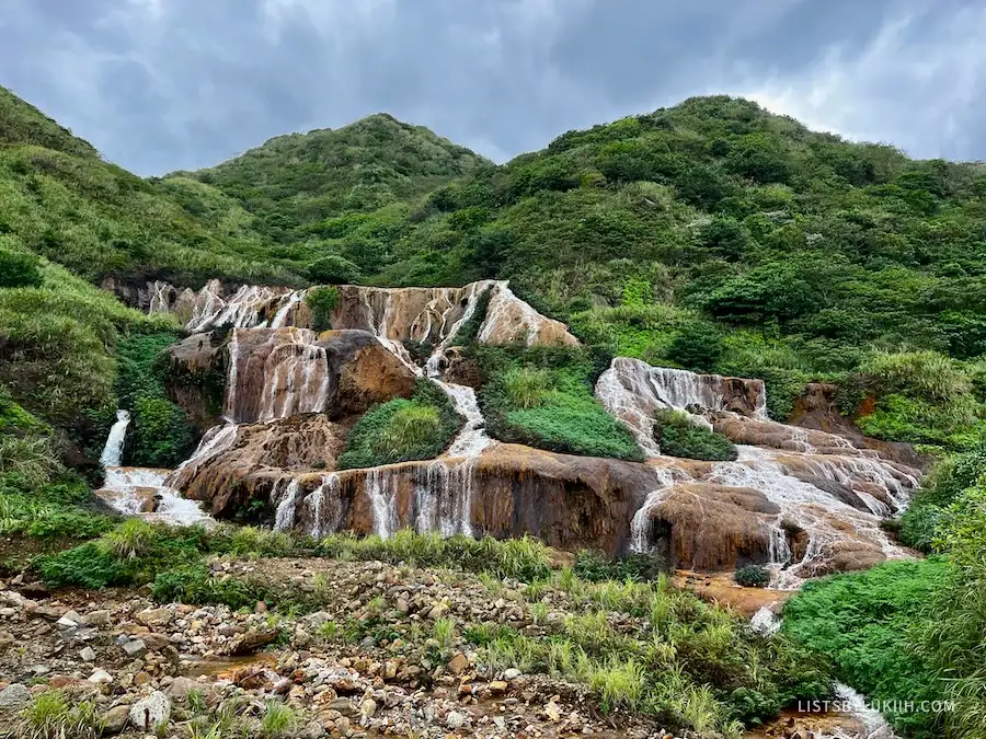 A short waterfall coming down a set of golden-colored rocks against a lush, green mountain.