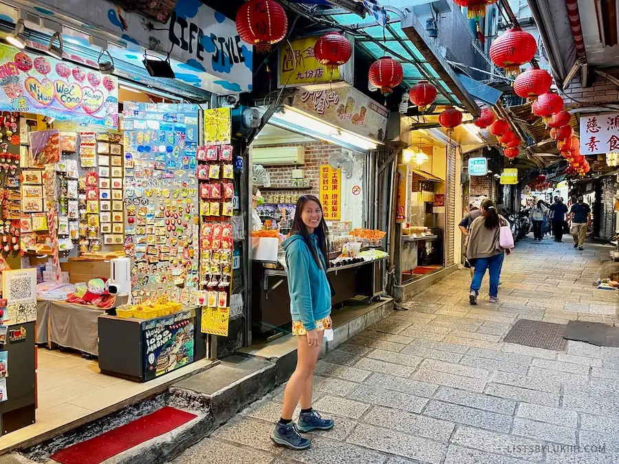 A woman smiling at the camera with red lanterns and small vendor shops in the background.