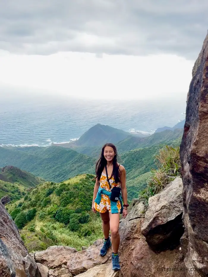 A woman standing at the edge of a mountain with the ocean in the background.