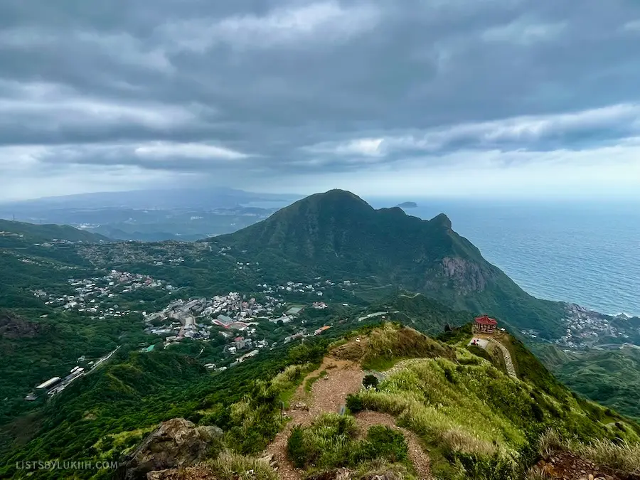 A view of a coastal town with a mountain in the middle and a shrine on a trail.