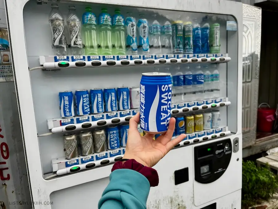 A hand holding up a blue can that says "Pocari Sweat" with a vending machine of Asian sport drinks in the background.