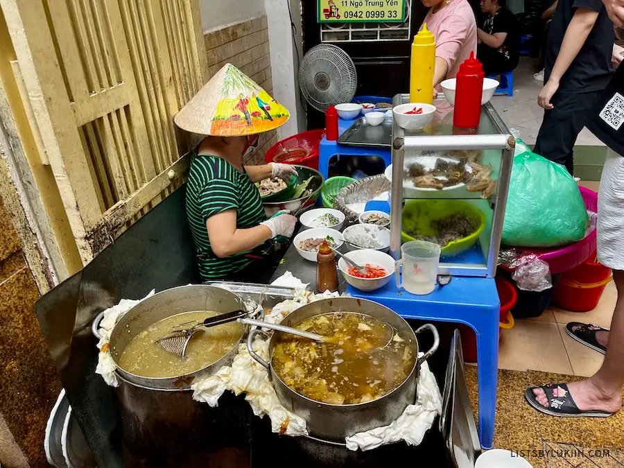 A woman wearing a straw hat cooking large pots of soup.