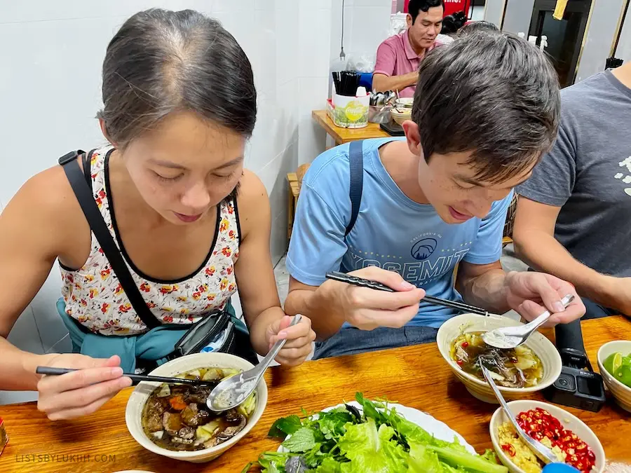 Two people sitting at a table eating noodle with a dark broth.