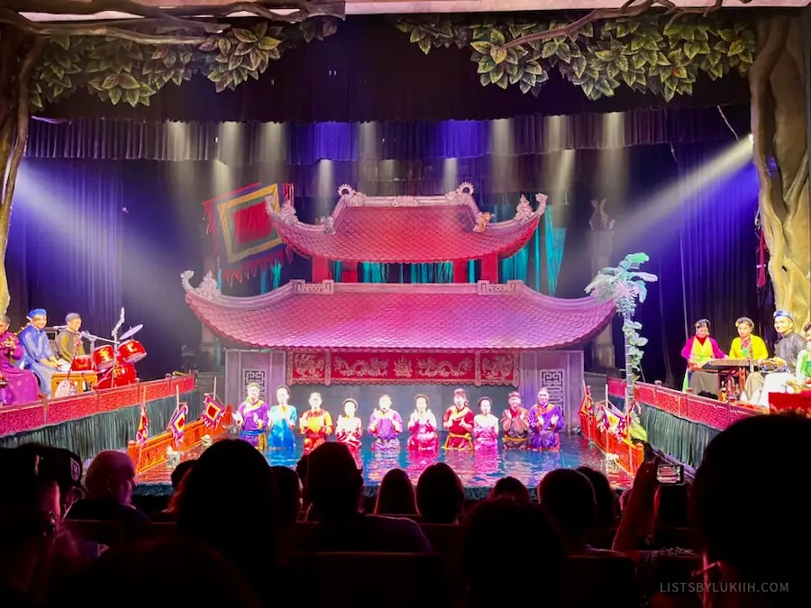 An indoor theater with an Asian-building prop and ten performers standing in water.