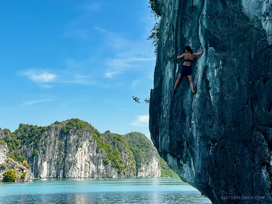 A woman climbing up a dark limestone rock over water.