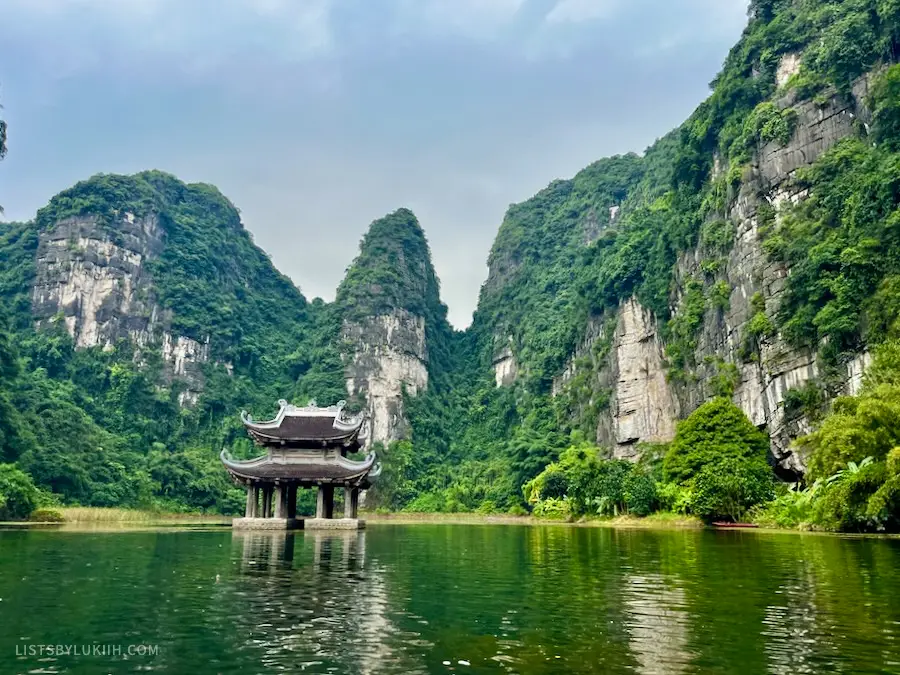 River with emerald water with a small temple inside the water and limestone mountains in the background.