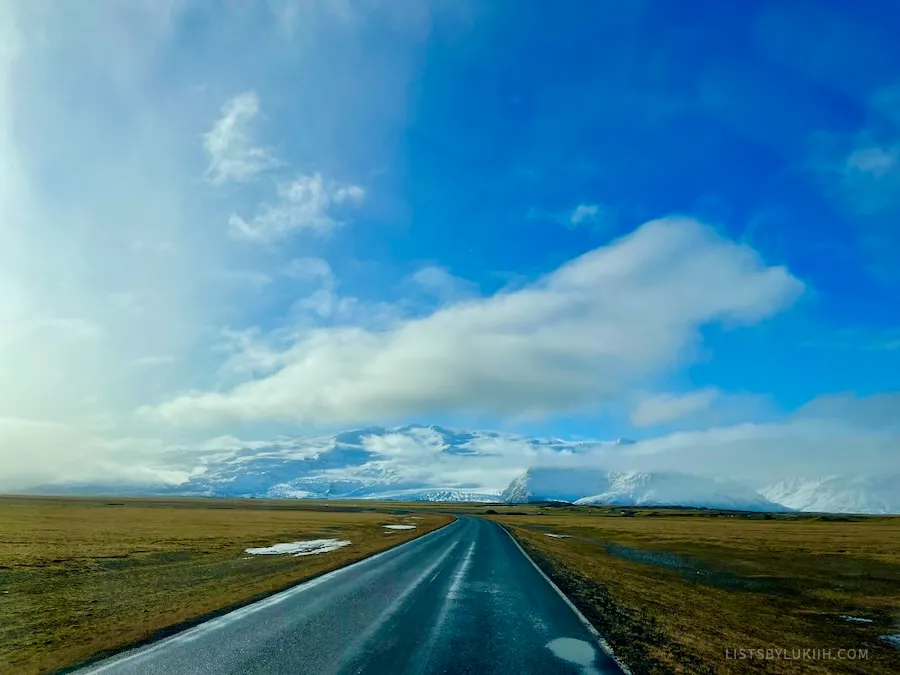 A road going through an empty landscape with glacier mountains in the background.