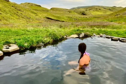 A woman sitting in natural water looking out at a green valley.