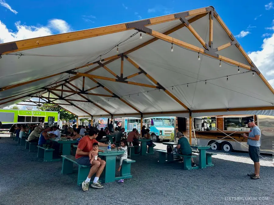 People sitting at several tables under a large canopy with food trucks on the side.