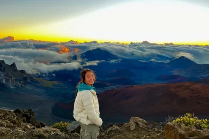 A woman standing on a mountain with clouds and a sunrise in the background.
