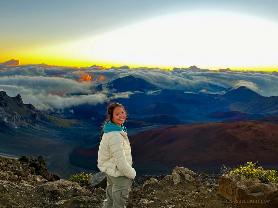 A woman standing on a mountain with clouds and a sunrise in the background.