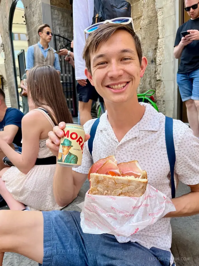 A man holding a sandwich with salami and an Italian beer.