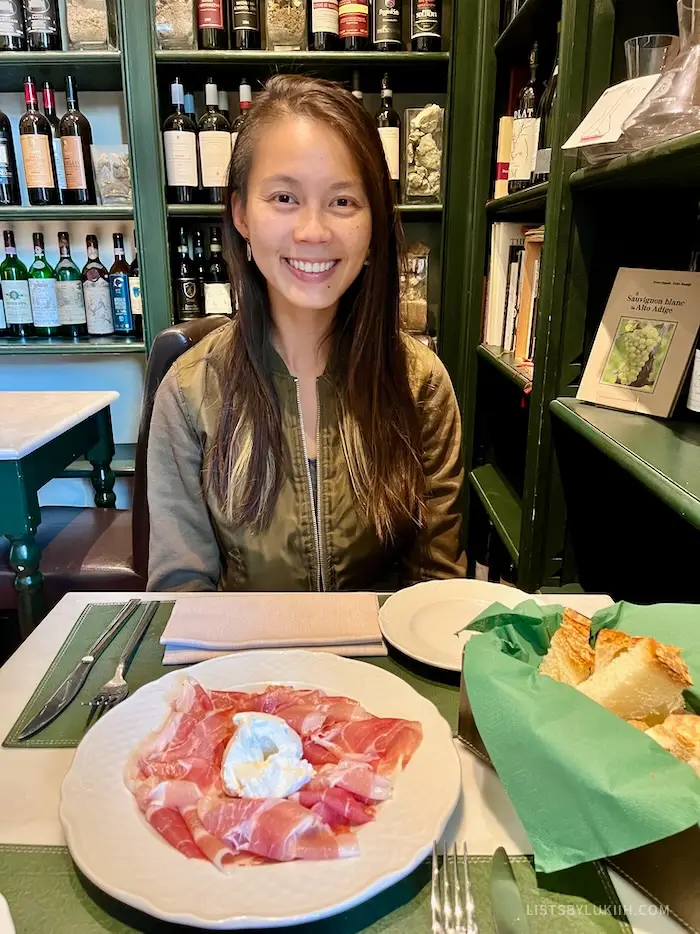 A woman sitting in front of a plate with of prosciutto with the walls decorated with wine bottles.
