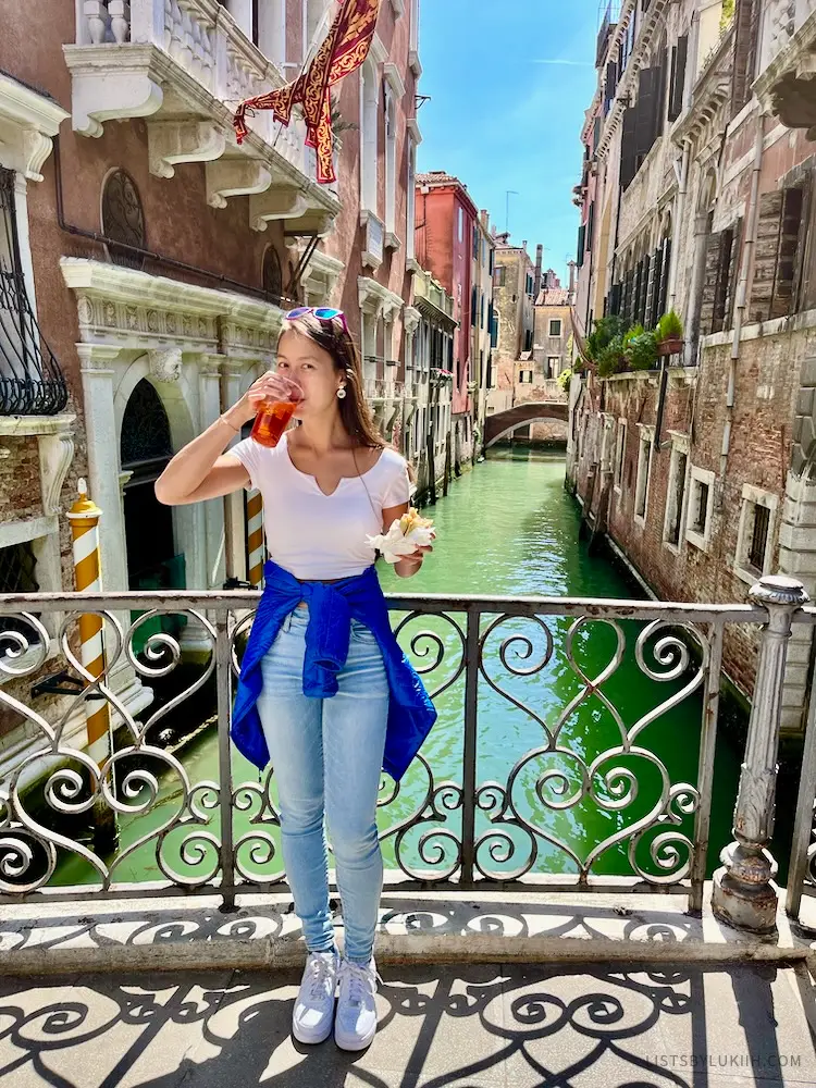 A woman holding a cup with red-orange liquid standing by a small, colorful canal.