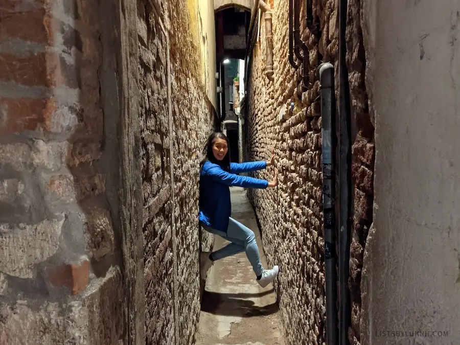 A woman in a very narrow street where she can touch both sides of the wall easily.